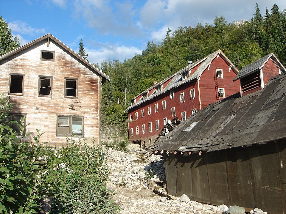 Weathered wooden building and red mill buildings at Kennecott, with a forested hillside behind