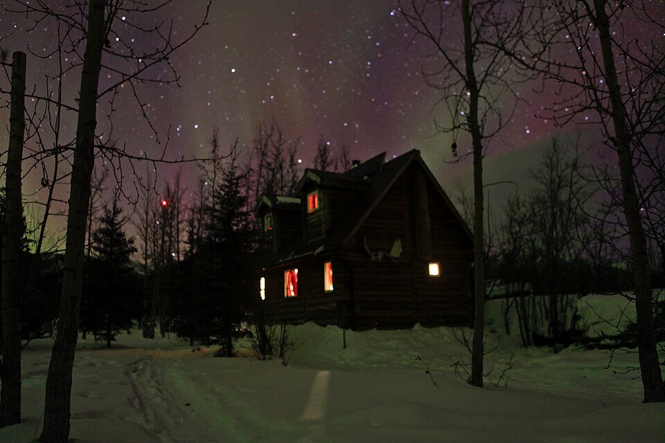 Cielo nocturno con una tenue aurora sobre una casa de madera con ventanas iluminadas en Wiseman, Alaska