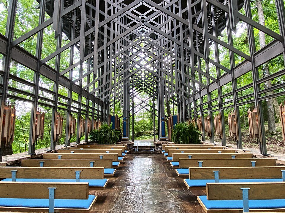 Interior of Thorncrown Chapel: wooden pews with blue cushions on a flagstone floor, under a lattice of beams and glass walls