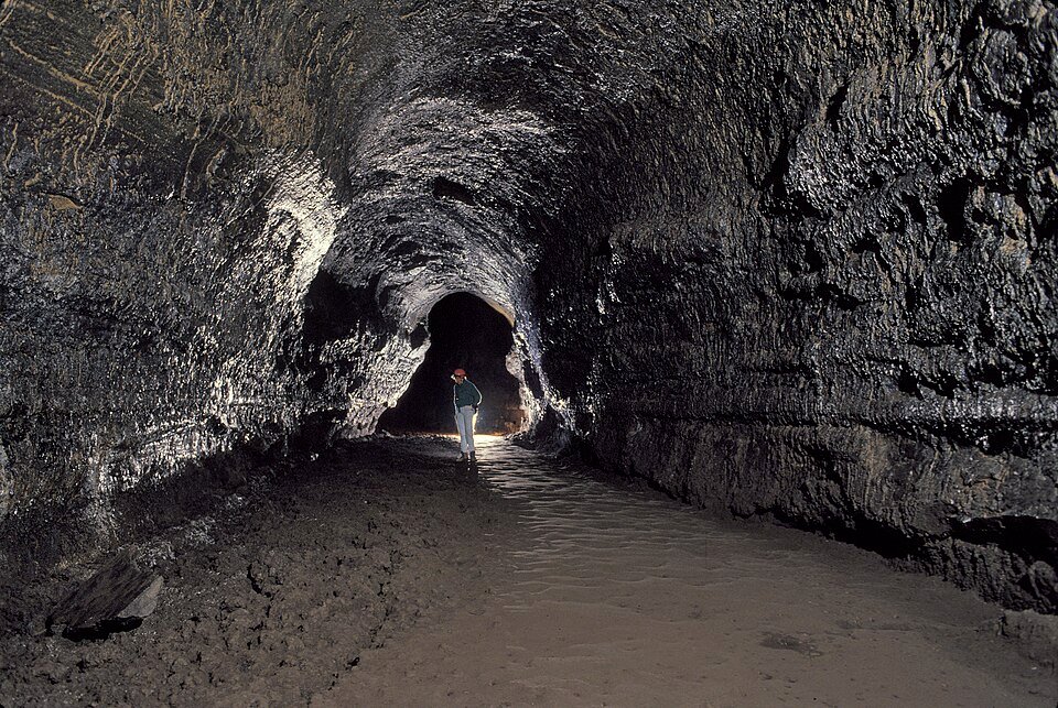 A wide lava tube passage with dark, glossy walls and a visitor standing in the distance