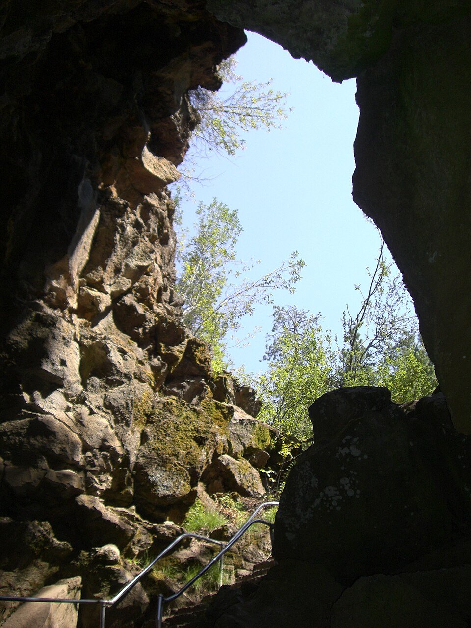Looking up from inside the collapsed entrance of Lava River Cave: dark rock walls, a handrail and trees against the sky