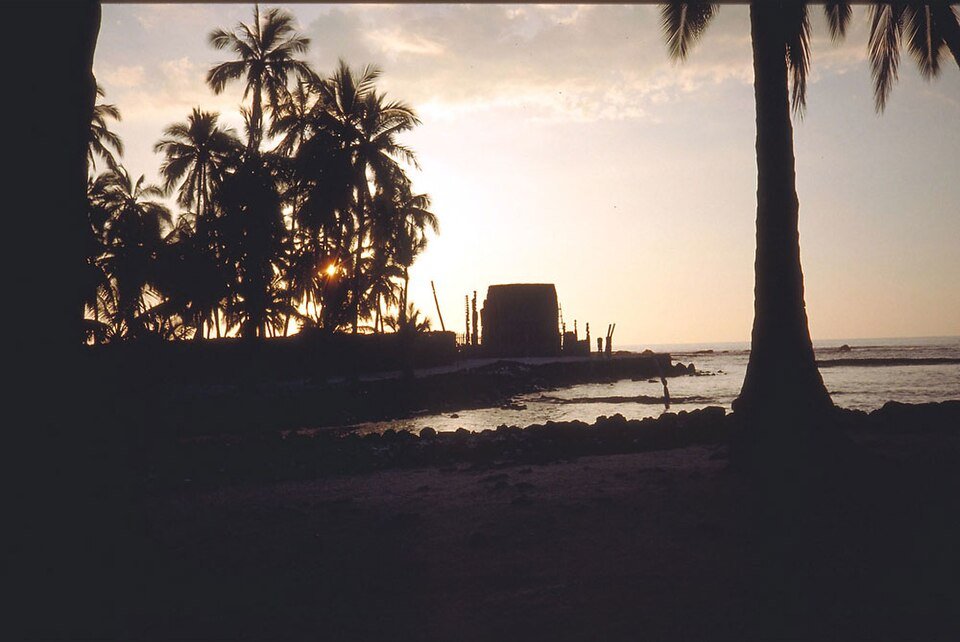 The thatched temple and carved figures in silhouette at sunset, with palms beside the shore