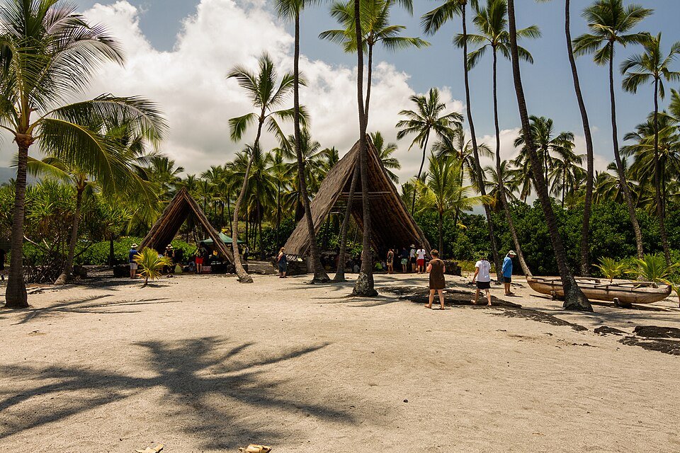 Thatched hale among palm trees on pale sand, with an outrigger canoe and a few visitors