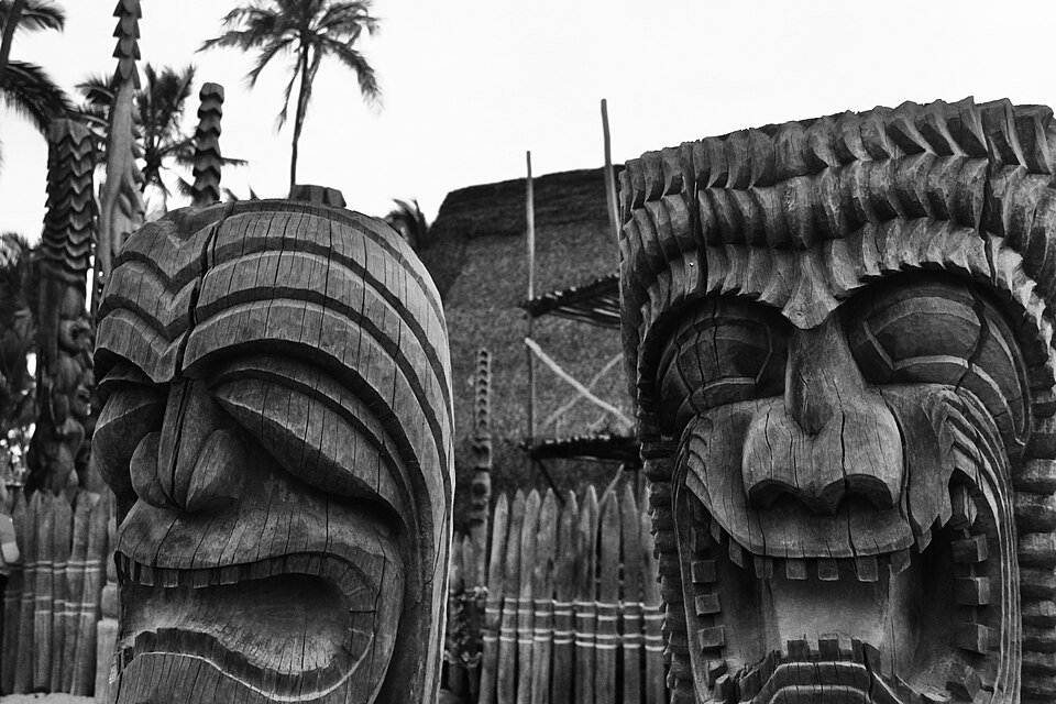 Close view of two large carved wooden figures, with a thatched structure and a wooden fence behind