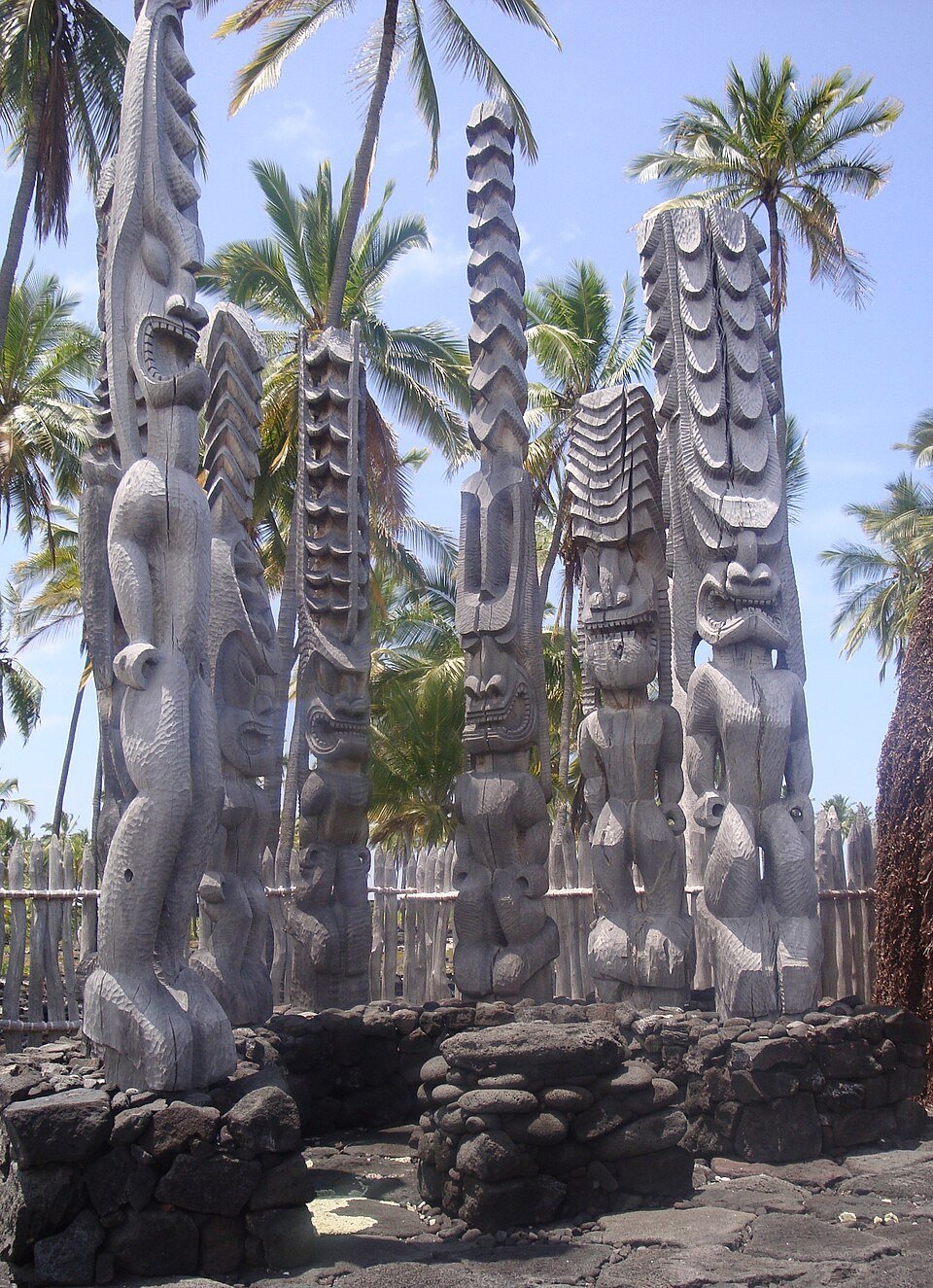 A group of tall carved wooden figures on a lava-rock platform among palm trees