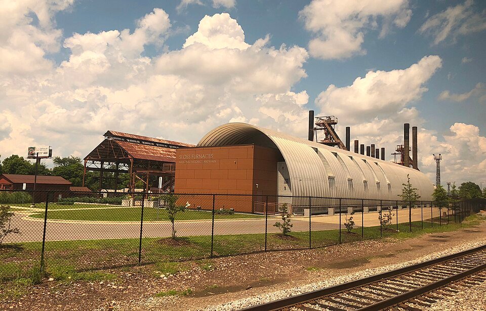 O edifício de teto arqueado com a placa da Sloss Furnaces, visto da linha do trem, com as chaminés atrás