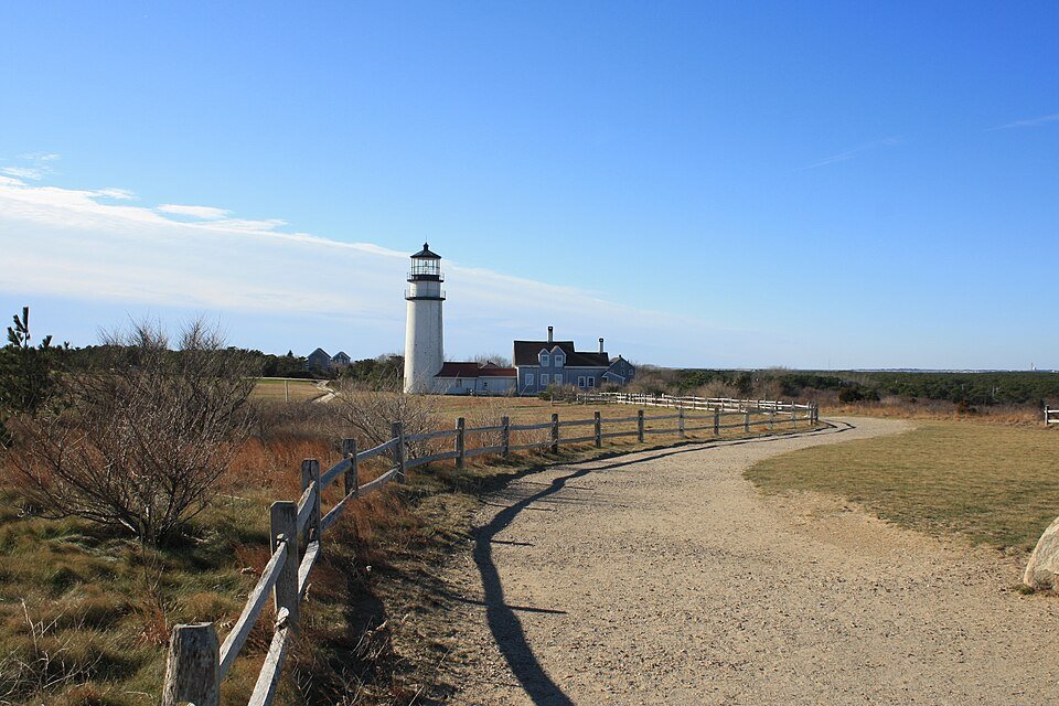 Highland Light y la casa del farero a lo lejos, vistos desde un camino de grava junto a una valla de madera