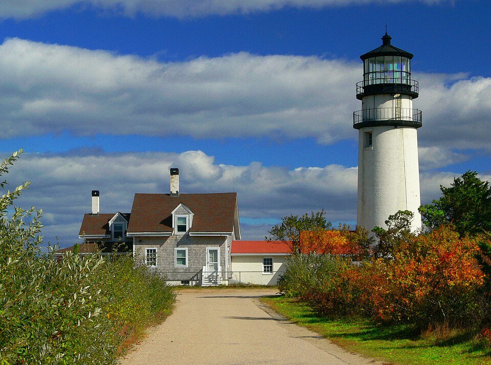Highland Light y la casa del farero junto a un camino de grava, con follaje otoñal y cielo azul