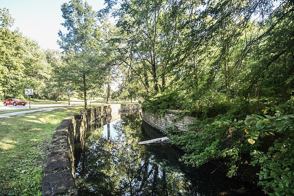 Um canal de água parada com muros de pedra ao lado de uma via do Rockefeller Park