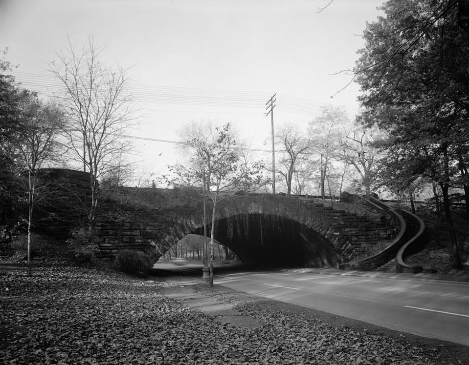 Uma vista antiga em preto e branco de uma ponte de pedra em arco sobre uma via do Rockefeller Park