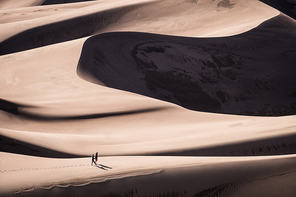 Wave-like sand dunes at Great Sand Dunes National Park, with two tiny hikers on the sand below