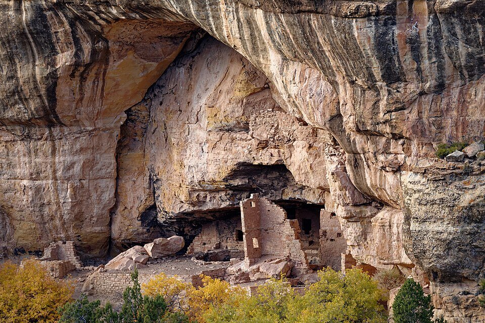 Stone ruins of a cliff dwelling tucked into an alcove under a sandstone overhang, with autumn foliage below