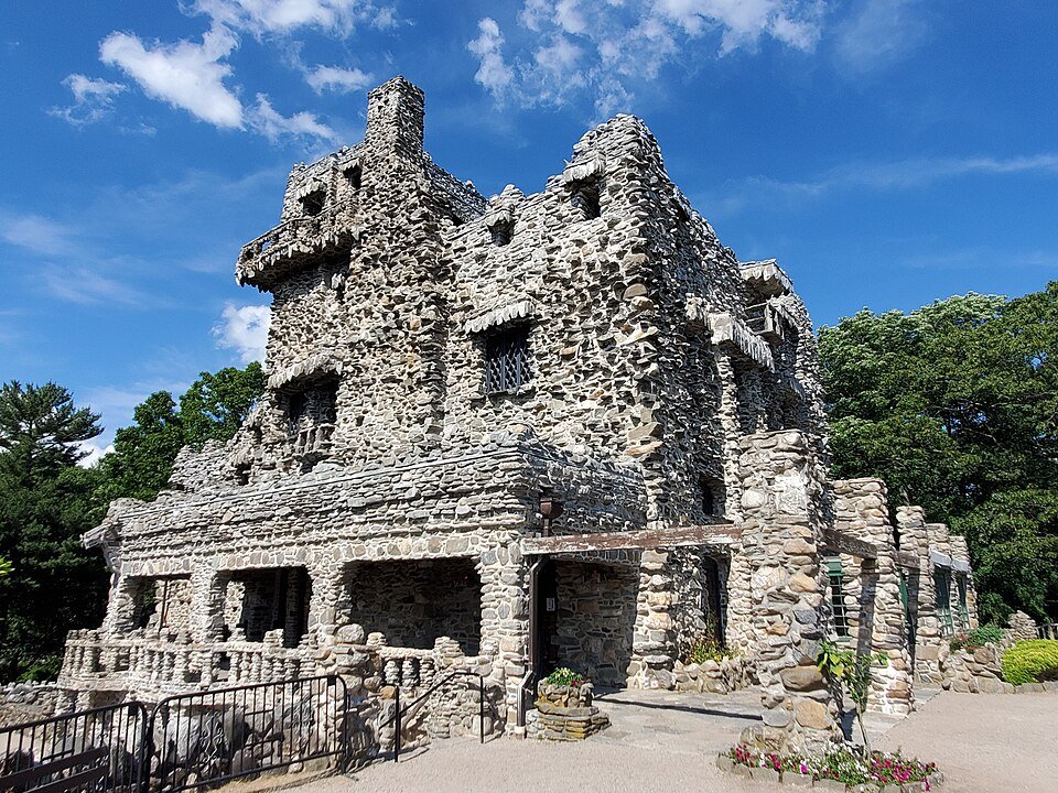 Rough stone walls, towers and covered porch of Gillette Castle against a blue sky, with flower beds in front