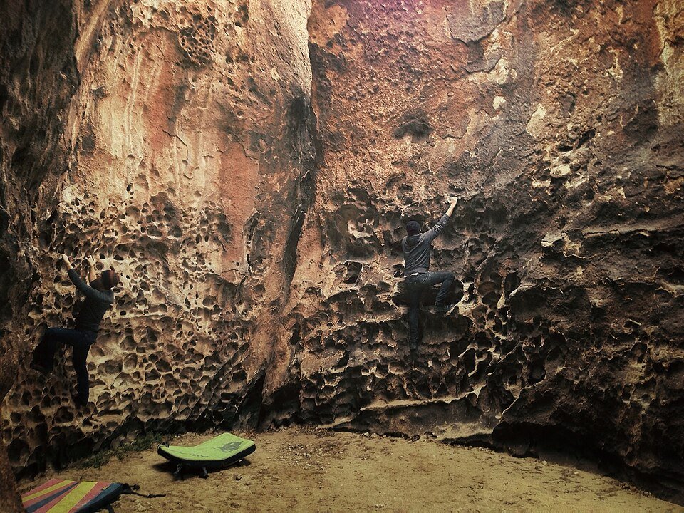 Two climbers on a rock wall full of small pockets and holes, with crash pads on the sandy ground