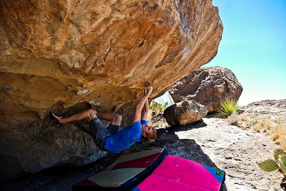 A climber hanging beneath a large overhanging boulder at Hueco Tanks, with yucca and prickly pear nearby