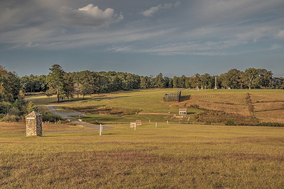 Open grounds of the site with a reconstructed section of stockade and monuments in the distance