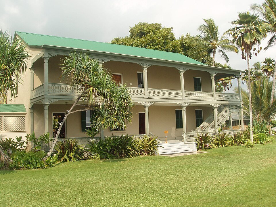 Parte trasera del Palacio Huliheʻe, un edificio de dos pisos con techo verde y galerías de madera, vista desde el césped