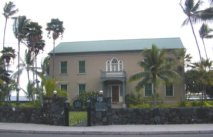 Fachada del Palacio Huliheʻe, un edificio de dos pisos con techo verde tras un muro de roca de lava negra y una reja de hierro, vista desde la calle