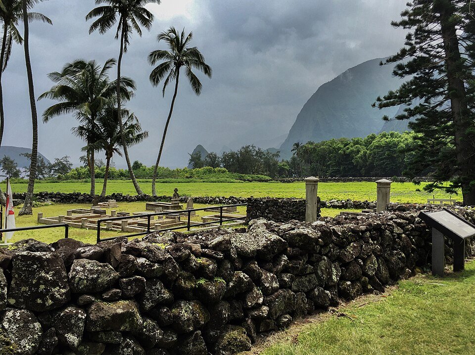A lava-rock wall and a walled enclosure with low stone structures among palm trees, with steep green cliffs behind, at Kalaupapa
