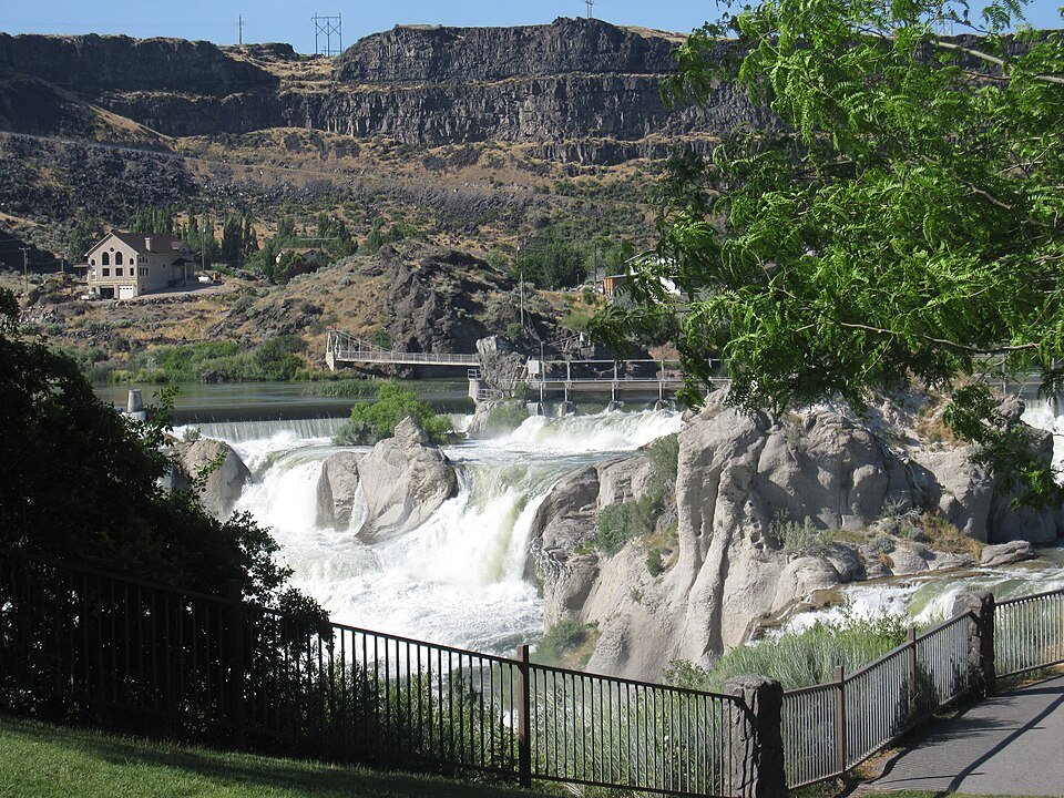 Shoshone Falls seen from a fenced viewpoint, with white water rushing over rock ledges below a footbridge