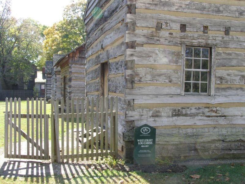 Log cabins along West Street in New Harmony, Indiana, behind a wooden gate and a marker