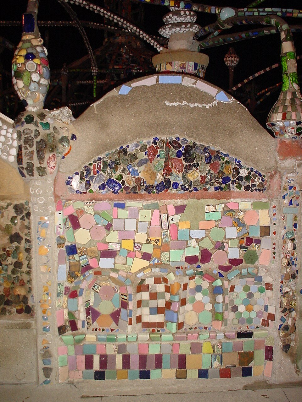 Mosaic of tile, stone and glass on a wall at the Watts Towers, with the towers behind at night