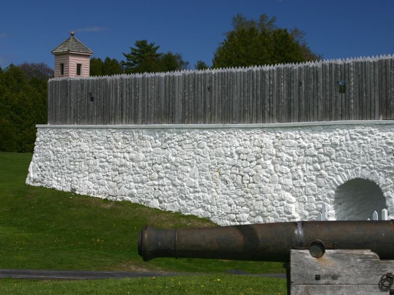 Whitewashed stone wall and wooden stockade of Fort Mackinac, with a cannon barrel in the foreground