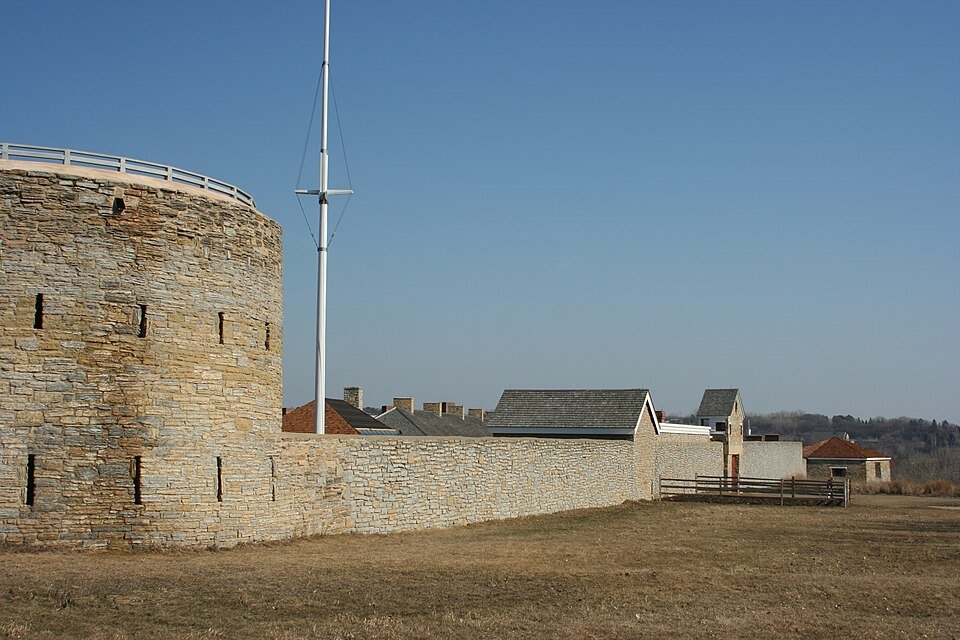 La torre redonda y un largo muro de piedra, con un asta de bandera y los edificios restaurados del fuerte al fondo