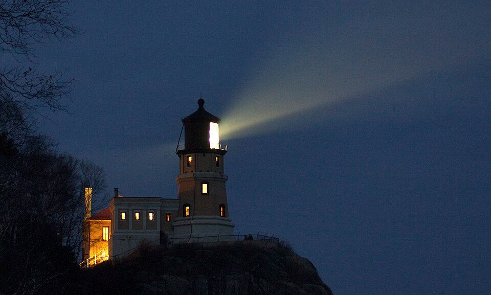 El faro Split Rock encendido al anochecer sobre su acantilado, con su haz de luz cruzando el cielo oscuro