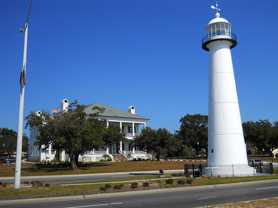 El faro de Biloxi, una torre blanca junto a una calle, con un edificio blanco de dos plantas detrás
