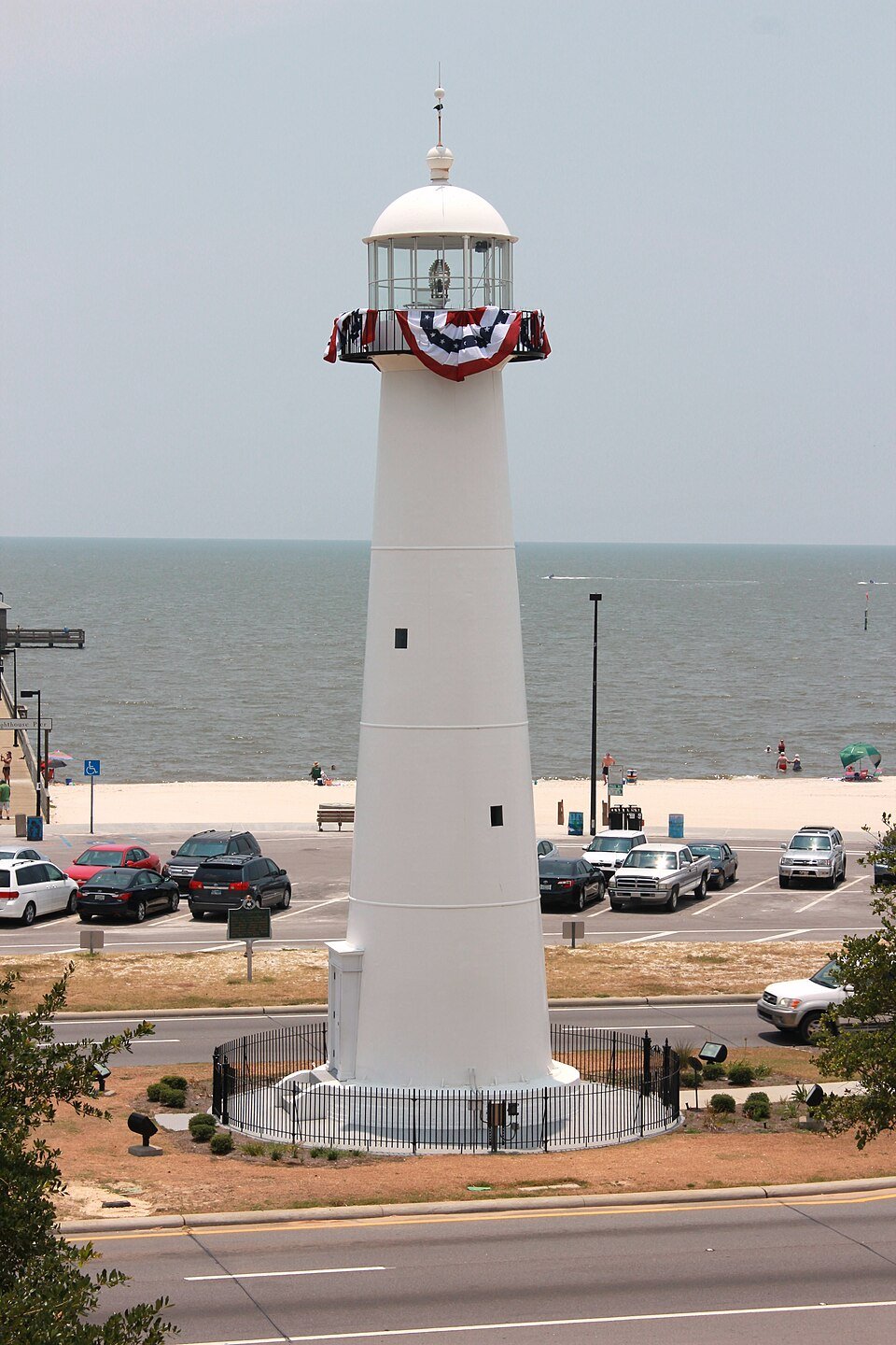 El faro de Biloxi visto desde arriba, con banderas decorativas en la galería y la playa y el Golfo detrás