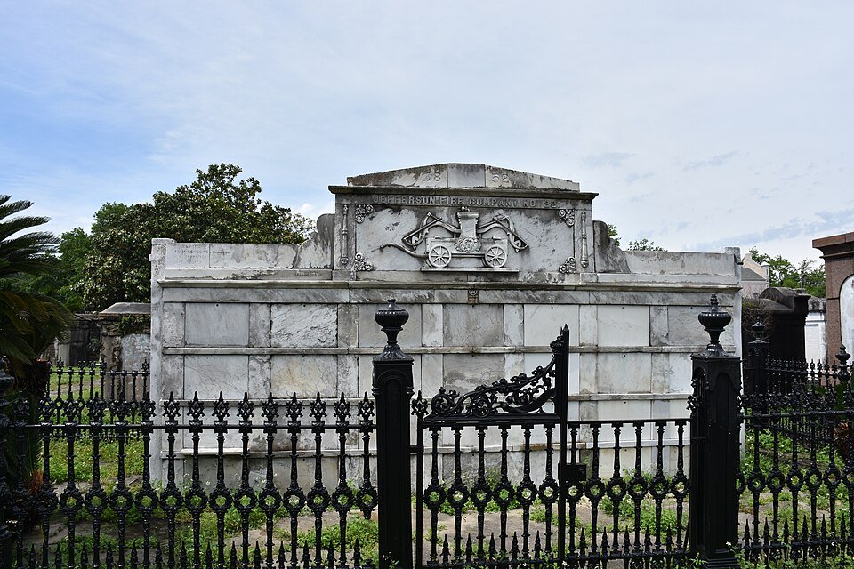 A tomb with a carved fire engine relief and a fire company name, behind an iron fence