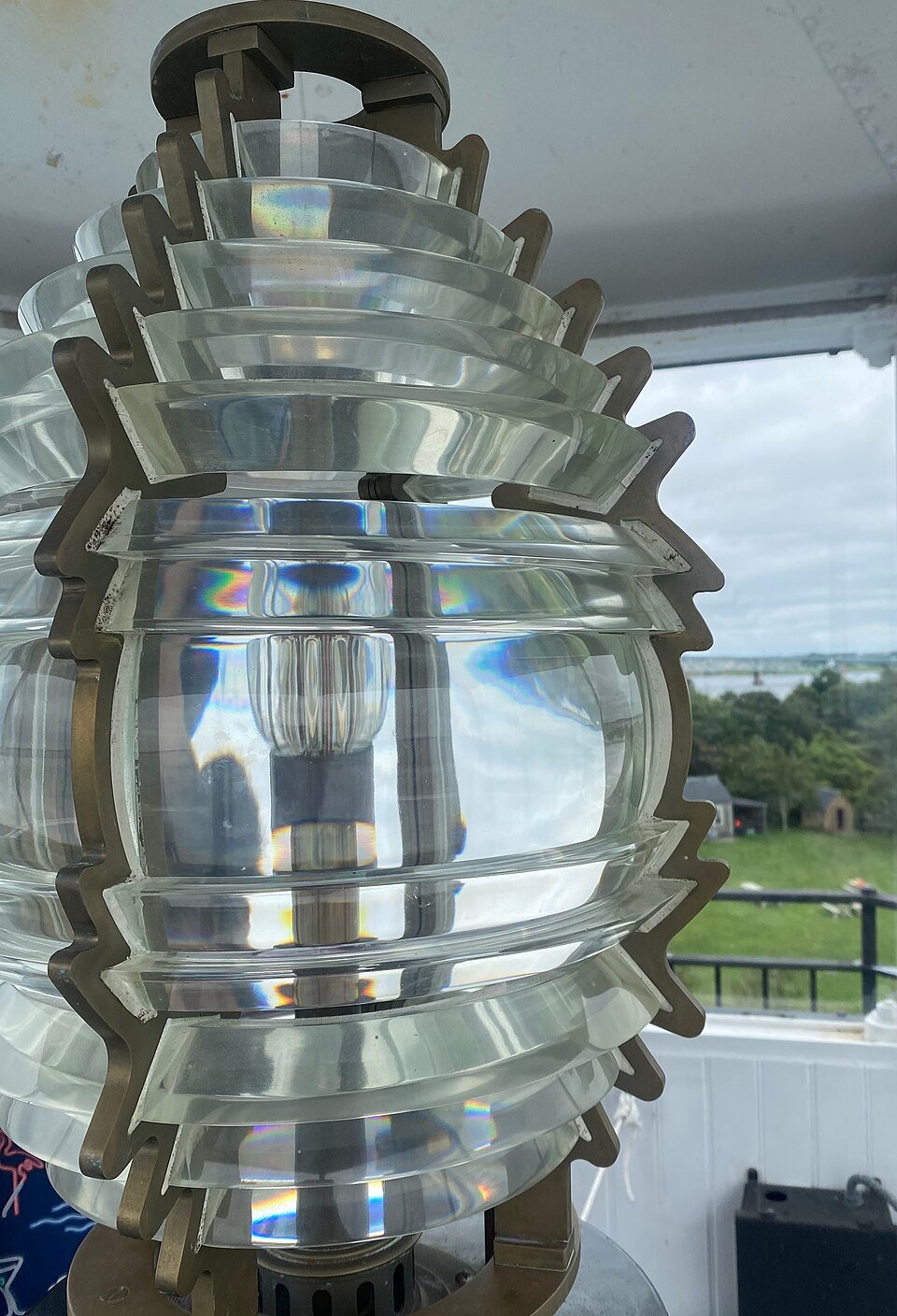 A Fresnel lens with ring-shaped glass prisms in a bronze frame, from Rose Island Light, with a view of the island through the window