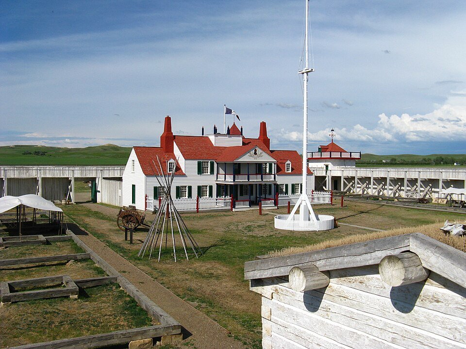 Dentro do Fort Union Trading Post reconstruído: uma casa branca de telhado vermelho, um mastro de bandeira e a estrutura de um tipi dentro de muros de paliçada caiados, com colinas de pradaria ao fundo