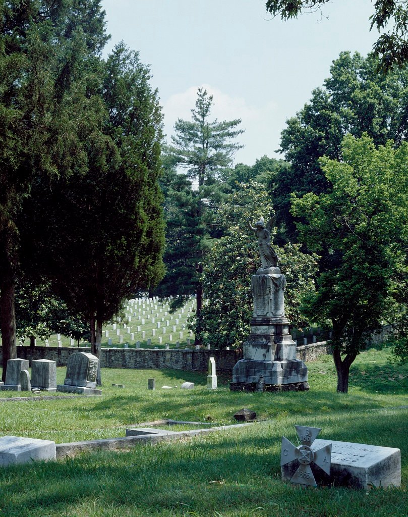 An angel monument and old gravestones, with rows of white headstones beyond a stone wall
