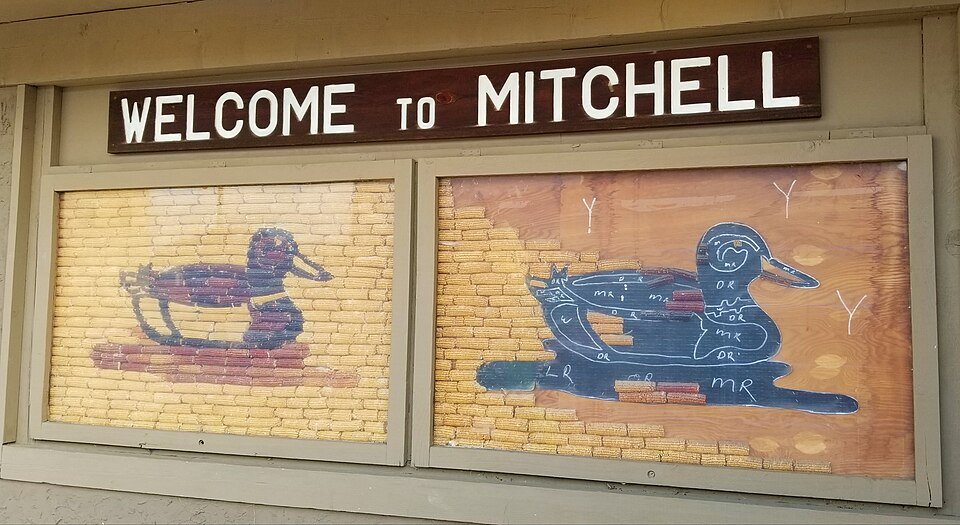 Painéis de murais de milho com patos ao lado de uma placa de boas-vindas a Mitchell, um deles ainda com marcas de giz como guia