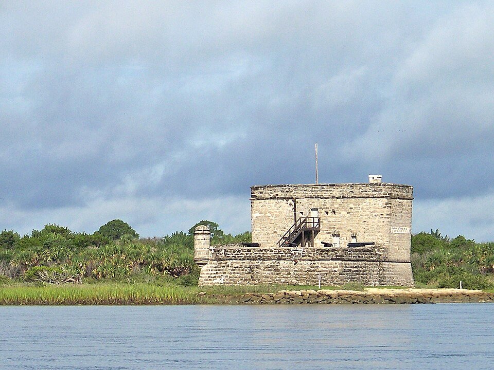 Fort Matanzas al otro lado del agua bajo un cielo de tormenta, con marisma y palmeras detrás