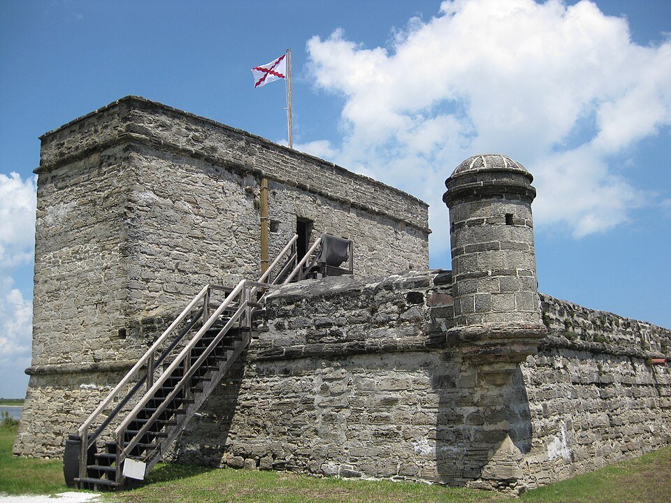 Vista cercana de la torre de coquina, su escalera de madera y una garita de esquina, con la bandera española