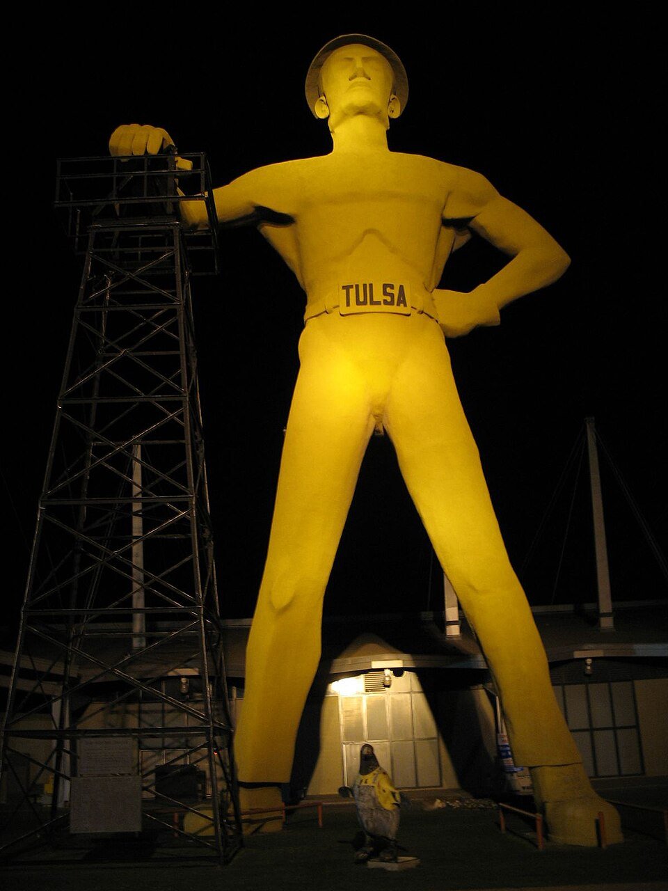The Golden Driller lit at night, with the derrick at its left and a small statue in the foreground