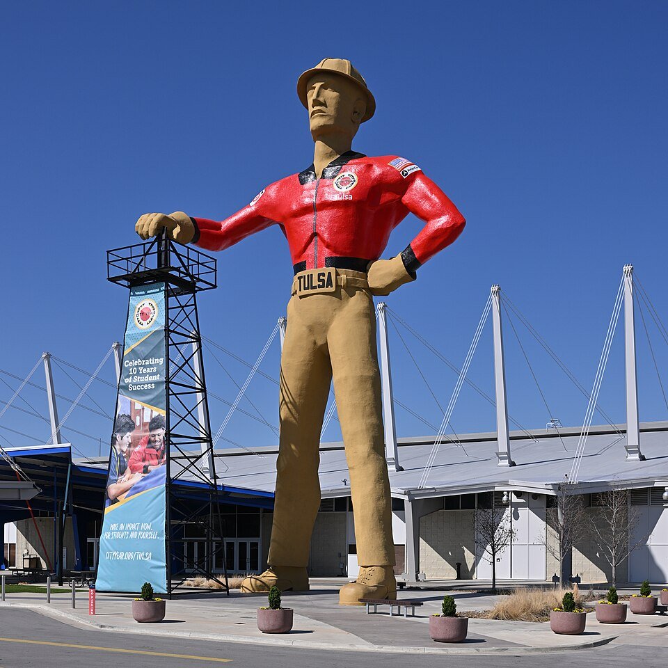 The Golden Driller in a red jacket and hard hat, one hand on the derrick, in front of the Expo Center