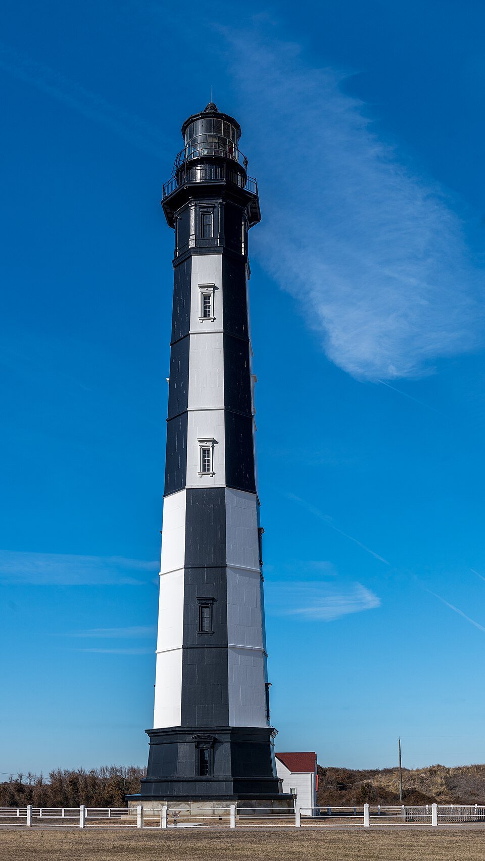 The black-and-white striped 1881 lighthouse against a blue sky, with a white fence and a small white building at its base