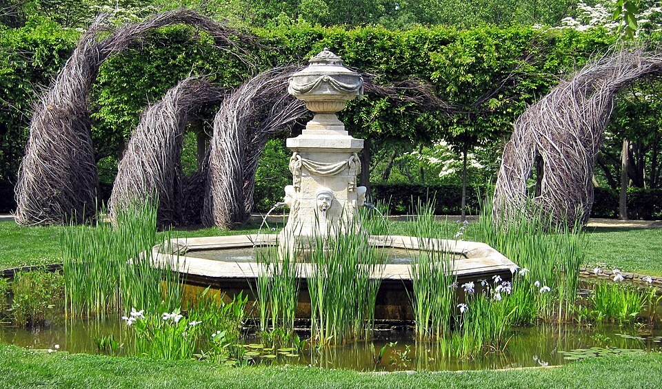 Stone fountain in a pool with irises, with woven twig arches behind, in the gardens of Dumbarton Oaks