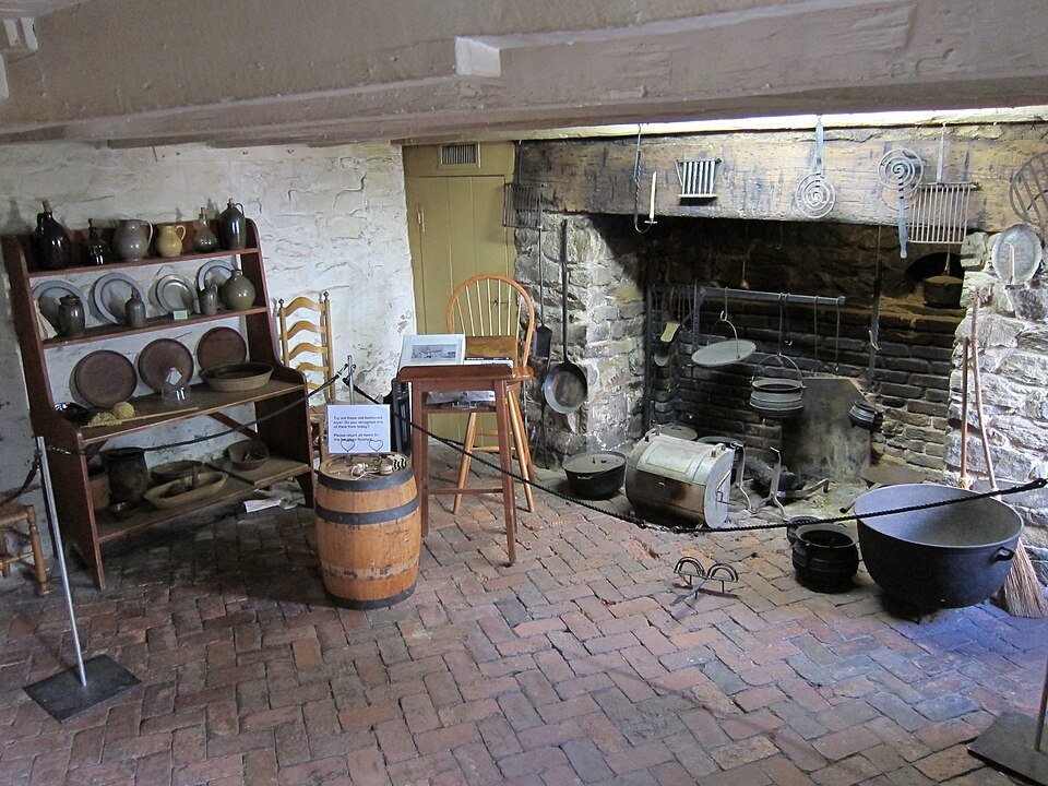Rear kitchen of the Old Stone House, with a large stone fireplace, iron pots and a brick floor