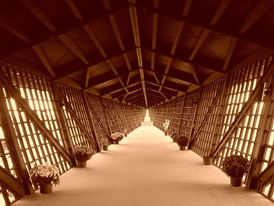 Vista ao longo da Infinity Room, um corredor longo e estreito com paredes inclinadas de treliça e vasos de flores no chão
