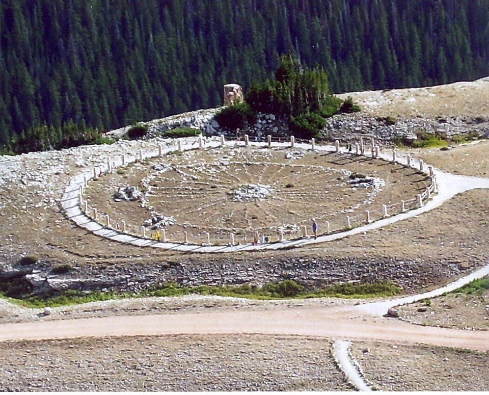 O Medicine Wheel visto de cima: um anel de pedras com raios dentro de uma cerca baixa e de uma passarela, em uma crista desnuda com floresta de coníferas ao fundo